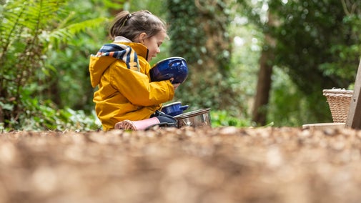 Young child in a yellow raincoat sitting on the forest floor among leaves playing with a saucepan